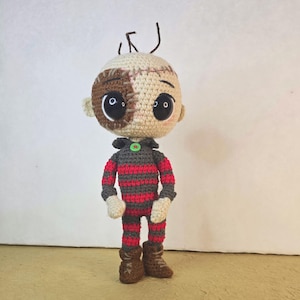 May include: A handmade crochet doll with a unique design. The doll features a stitched head with a brown patch, large black eyes, and brown hair. It wears a red and gray striped outfit with brown boots. The doll has a green button on its collar.