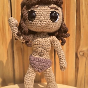 May include: A handmade crochet doll with brown hair and large black eyes. The doll is wearing a purple bottom and has a tan skin tone. The doll is standing with one arm raised. The doll is made of yarn.