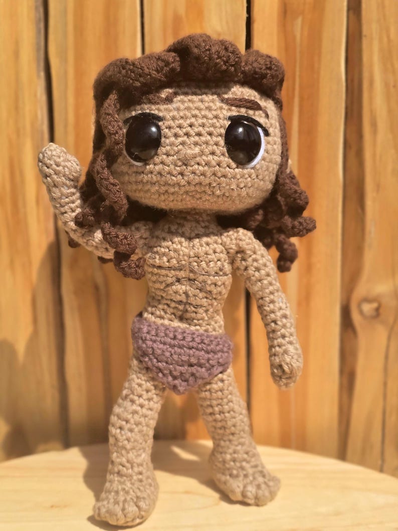 May include: A handmade crochet doll with brown hair and large black eyes. The doll is wearing a purple bottom and has a tan skin tone. The doll is standing with one arm raised. The doll is made of yarn.