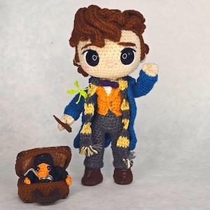 May include: A crocheted doll with brown hair, large black eyes, wearing a blue coat, orange vest, and a yellow and black scarf. The doll holds a brown wand and a small green creature. Beside it sits a brown treasure chest containing a black creature.