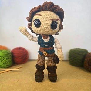 Puede incluir: Muñeca de ganchillo hecha a mano que representa a un personaje con piel bronceada, cabello castaño y ojos negros grandes. La muñeca viste un chaleco verde azulado, pantalones marrones y botas. Una pequeña bolsa cuelga del cinturón.