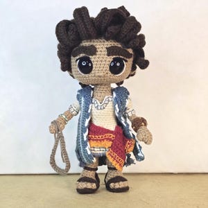 May include: A handmade crochet doll with brown skin, dark brown curly hair, and large black eyes. The doll wears a white shirt, a blue vest, and a red and orange skirt. It holds a rope in its hand and wears sandals.