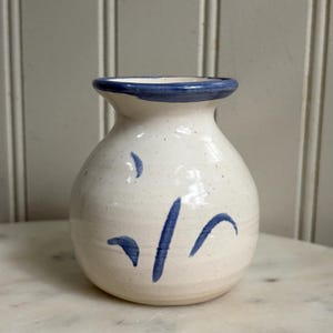 May include: A small, round, off-white ceramic vase with a speckled finish. The vase features a flared rim painted with a solid blue band and simple, hand-painted blue brushstroke markings scattered across the body of the vessel.
