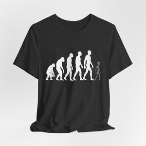 May include: Dark gray t-shirt featuring a white graphic depicting the evolution of humans, culminating in an alien figure. The design shows a progression from ape-like to human forms, with the alien at the end. The shirt is made of a soft material.