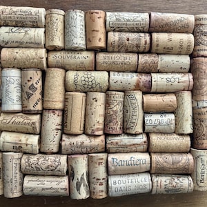 May include: A collection of used wine corks, arranged in a rectangular pattern. The corks display various brand names and logos, with a mix of light and dark brown tones. Some corks have text like "Columbia Crest" and "Bandiera".