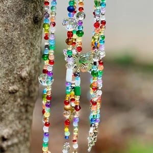 May include: A decorative wind chime featuring three hanging strands of multi-colored glass and plastic beads suspended from a horizontal wooden piece. The strands include various shapes, sizes, and colors of beads, a central butterfly charm, and small brass bells.