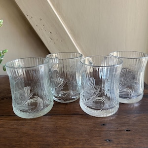 May include: A set of four clear glass tumblers featuring a vertical ribbed texture along the upper rim and a molded floral pattern around the base. Each glass has a slightly curved silhouette and a smooth, transparent finish throughout the body.