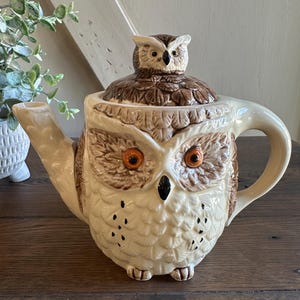 May include: A ceramic owl-shaped teapot with a cream-colored body and brown and tan details. The teapot features a detailed owl face with orange eyes and a small owl figurine on the lid. The handle and spout are curved.