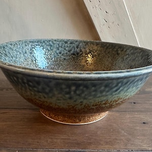 May include: A large, handcrafted ceramic bowl with a textured, speckled surface. The bowl features a gradient of colors, including shades of blue, green, and brown. It has a wide rim and sits on a small, round base, perfect for serving.
