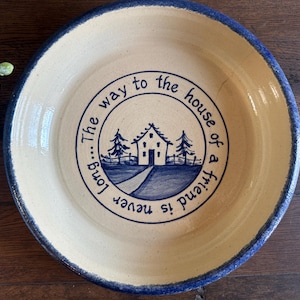 May include: A round, cream-colored ceramic plate with a blue rim. The center features a blue illustration of a house, trees, and a path, encircled by the text "The way to the house of a friend is never long..."