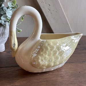 May include: A ceramic swan-shaped creamer with a curved neck and a yellow body. The swan's head and neck are a creamy white, and the body has a textured, feathered design. The creamer sits on a wooden surface.