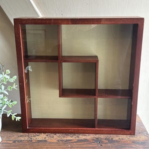 May include: A square, dark-stained wooden display case with a glass front and multiple interior shelves. The case has a warm, reddish-brown hue and is designed to showcase small items. The background is a light, neutral color.