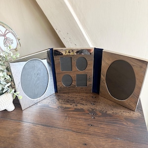 May include: A silver-toned metal triptych photo frame featuring three hinged panels. The two outer panels contain large oval openings, while the center panel features four smaller cutouts consisting of two rectangles and two ovals for photos.