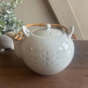 May include: A white ceramic teapot with a floral design and a bamboo handle. The teapot has a rounded shape and a small spout. The handle is curved and attached to the top of the teapot. The teapot is sitting on a wooden surface.