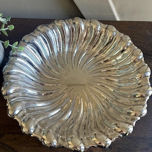 May include: A silver-colored, decorative serving dish with a fluted, radial design. The dish has a scalloped edge and a reflective surface, suitable for displaying food or as a decorative accent. The dish is round and sits on a dark wooden surface.