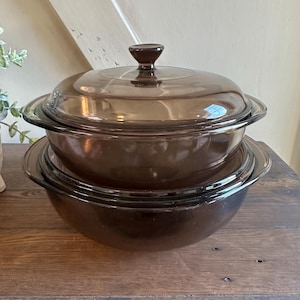 May include: Two amber-colored glass casserole dishes stacked on a wooden surface. The top dish has a matching lid with a brown knob. The dishes are round with a slightly flared rim, suitable for baking and serving.