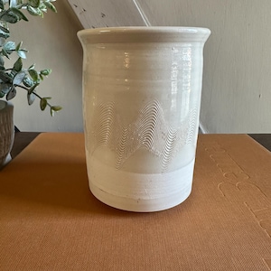May include: A cream-colored ceramic utensil holder with a wavy, textured design. The cylindrical holder has a slightly flared rim and sits on a brown surface. The design is a series of horizontal lines that create a wave pattern.