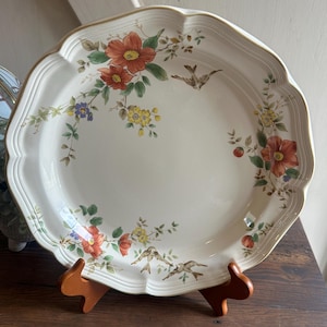 May include: A large, off-white ceramic serving platter with a scalloped edge and gold trim. The platter is decorated with floral and bird motifs in shades of red, yellow, and green. The platter is displayed on a wooden stand.