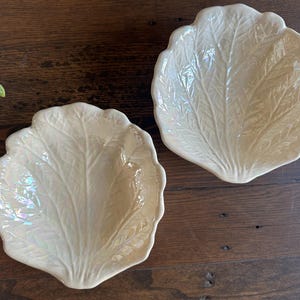 May include: Two off-white, leaf-shaped ceramic serving dishes. The dishes have a pearlescent finish and detailed veining. They are placed on a dark wooden surface, suggesting a decorative or serving purpose.