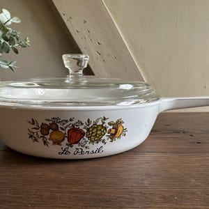 May include: White casserole dish with a clear glass lid and handle. The dish features a colorful vegetable design with the text "Le Persil". The dish is on a wooden surface.