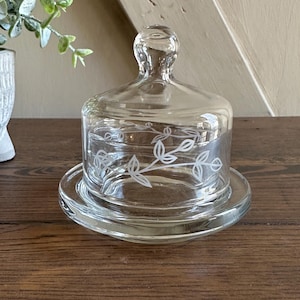 May include: A clear glass butter dish with a lid. The dish has a round base and a domed lid with a small knob. A delicate floral design is etched around the sides of the dish. The dish is sitting on a wooden surface.