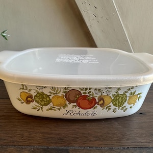 May include: A rectangular, off-white casserole dish with a matching lid. The dish features a colorful vegetable design with the word "L'Echalote" printed on the side. The lid has a handle on each side.
