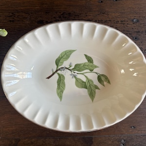 May include: Oval serving dish with a cream-colored base and a gold rim. The dish features a green and brown botanical design of leaves and small white flowers. The rim has a scalloped edge.