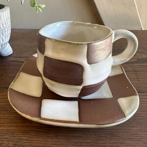 May include: A ceramic cup and matching square saucer featuring a hand-painted brown and off-white checkerboard pattern. The cup has a rounded body with a curved handle, and both pieces exhibit a glossy finish with slightly irregular, organic edges.