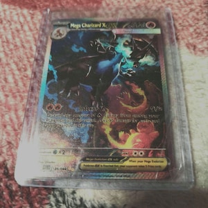 Puede incluir: Una carta coleccionable Pokémon Mega Charizard X EX en una funda protectora. La carta presenta una ilustración detallada de Charizard en tonos azules, negros y naranjas. La carta tiene el texto "Inferno X" y "Mega Evolution EX rule".
