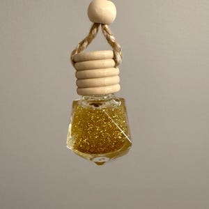 May include: A small, clear glass bottle filled with gold glitter, suspended by a tan and gold rope and a wooden bead. The bottle has a wooden cap with several stacked rings. The bottle is hanging against a neutral background.