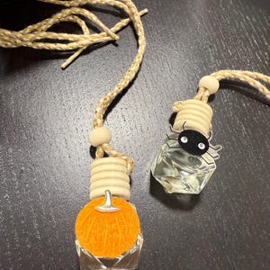 May include: Two car air fresheners. One is shaped like an orange pumpkin with a silver stem. The other is a clear glass bottle with a black spider decoration. Both have a wooden bead and a gold and beige rope.