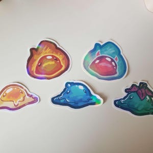 May include: Set of five colorful, holographic stickers featuring cartoon slime creatures in various shapes and colors. The stickers include orange, blue, and green slimes, with a glossy finish. Great for decorating laptops, notebooks, or water bottles.