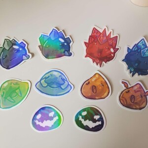 May include: A collection of colorful, cartoonish stickers featuring various crystal and slime designs. The stickers showcase a range of colors, including blue, green, red, and orange, with some having a holographic effect. The designs include spiky crystals and blob-like slimes.