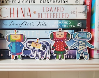 May include: Four colorful stickers featuring cartoon characters, including a robot, a cow, a strawberry person, and a mummy-like figure. The stickers are arranged in front of a stack of books with titles like "CHINA" and "The Daughter's Tale."