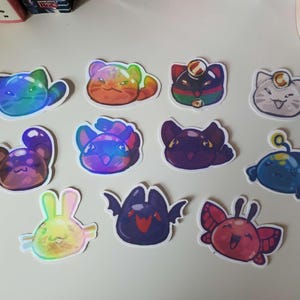 May include: A collection of colorful, holographic stickers featuring various cartoon creatures. Designs include cats, bunnies, and other whimsical characters in shades of blue, purple, orange, and red. Each sticker has a white border.