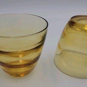 May include: Two amber-colored glass drinking glasses. One glass is upright, and the other is tilted on its side. The glasses have a conical shape with horizontal bands of darker amber color. The glasses are likely for serving beverages.
