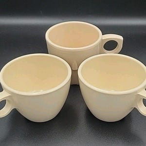 May include: Three cream-colored ceramic coffee cups with a rounded shape and a unique handle design. The cups are arranged on a dark surface, showcasing their simple yet elegant form. The interior of the cups has a lighter cream color.