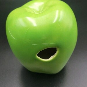 May include: A glossy, green ceramic apple with a hole on the side. The apple is a solid, vibrant green color and has a smooth, reflective surface. The hole is dark and oval-shaped. The apple is a decorative item.