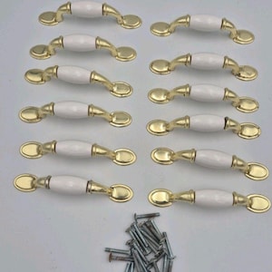 May include: A set of twelve cabinet pulls featuring a smooth white ceramic center handle with polished gold-tone metal ends and oval mounting plates. A collection of silver-colored metal screws is included at the bottom of the arrangement.
