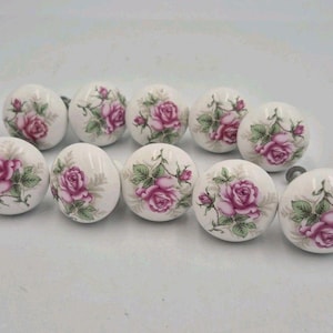 May include: A collection of ten round, white ceramic cabinet knobs, each featuring a detailed floral pattern of pink roses with green leaves and stems. The knobs are arranged in a group, showcasing a smooth, glossy finish and a consistent design.