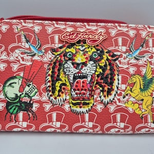 May include: A red rectangular wallet featuring an Ed Hardy logo, a central tiger head embellished with colorful rhinestones, and a repeating white pattern of skulls wearing top hats. The design includes illustrations of a green skull, a blue bird, and a yellow winged horse.