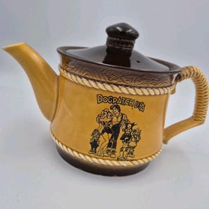 May include: A ceramic teapot with a tan body and a dark brown lid and base. The teapot features a rope-like texture around the top and bottom edges and a handle shaped like rope. It displays a black graphic of characters and the text DOGPATCH USA.