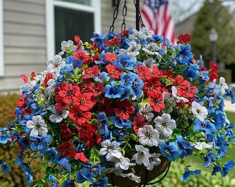 May include: A hanging basket filled with artificial flowers in red, white, and blue. The arrangement features small, five-petaled blossoms with green foliage, suspended by a black metal chain and hook from a tall, thin black metal stand.