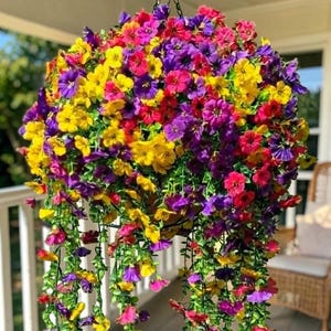 May include: A hanging basket filled with a dense arrangement of artificial flowers in vibrant shades of yellow, purple, and pink. The floral display features numerous small blossoms and green trailing stems suspended by a black metal chain and hook.