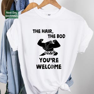May include: White t-shirt with a black graphic of a muscular figure flexing, with the text "THE HAIR, THE BOD, YOU'RE WELCOME" in black. The shirt is hanging on a wooden hanger.