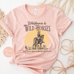 May include: Heather peach-colored t-shirt with a graphic design. The design features a person riding a horse in front of a yellow sun, with the text "Wildflowers & Wild Horses" and "FOUR FIFTHS OF RECKLESS AND ONE FIFTH OF JACK".