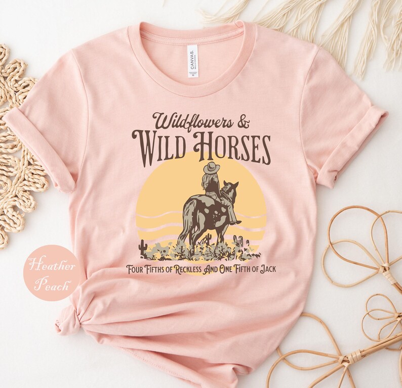 May include: Heather peach-colored t-shirt with a graphic design. The design features a person riding a horse in front of a yellow sun, with the text "Wildflowers & Wild Horses" and "FOUR FIFTHS OF RECKLESS AND ONE FIFTH OF JACK".