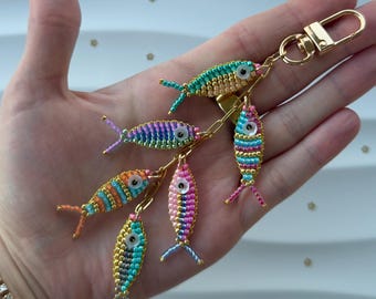 Puede incluir: Un llavero de pez con cuentas de colores con seis peces en varios colores, incluyendo azul, verde, naranja, rosa y morado. Los peces están unidos a una cadena y un cierre de color dorado. El llavero se sostiene en una mano.