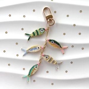 May include: A beaded fish keychain with a gold-colored clasp and chain. The keychain features five colorful fish with detailed beadwork in shades of green, blue, yellow, and white. Small gold star-shaped embellishments are scattered around the keychain.