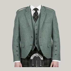 Puede incluir: Un traje de tres piezas de tweed verde claro con chaleco a juego y corbata a cuadros negros y grises. La chaqueta tiene dos bolsillos delanteros y botones decorativos en las mangas. El traje se combina con un kilt escocés tradicional.